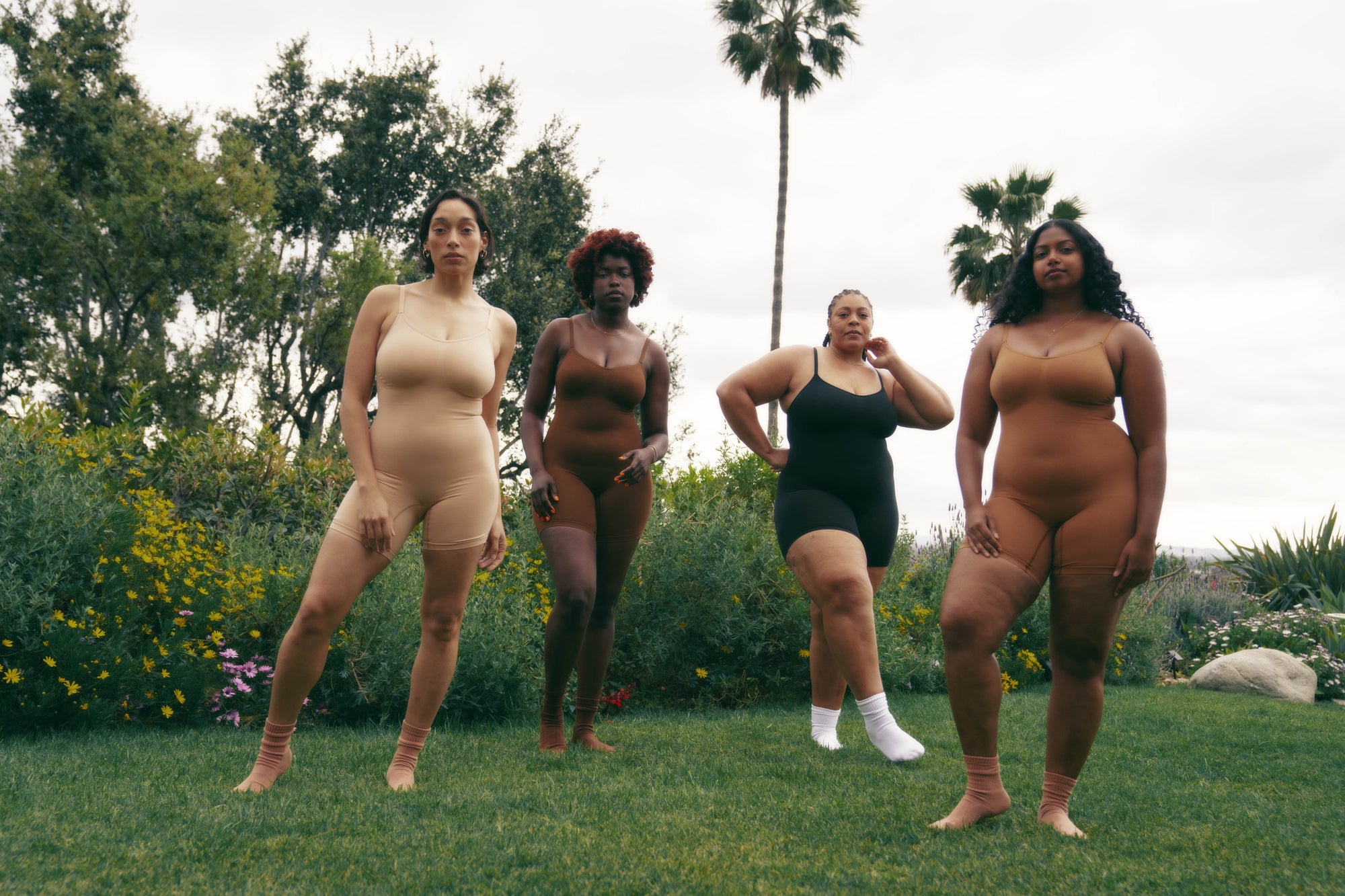 four models stand on a green field with lush tropical foliage around, wearing plant-based shapwear bodysuits in inclusive nude tones.