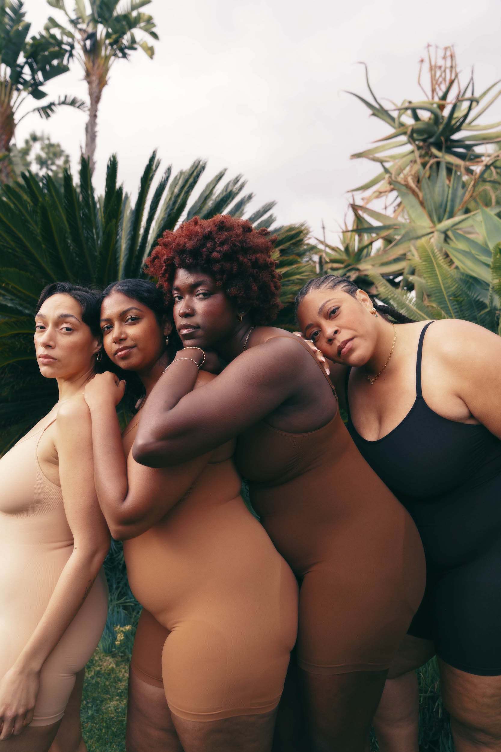 models in our plant-based sustainable shapewear leaning into each other as they look at the camera. light nude, medium nude, deep nude and black mid-thigh bodysuits are worn